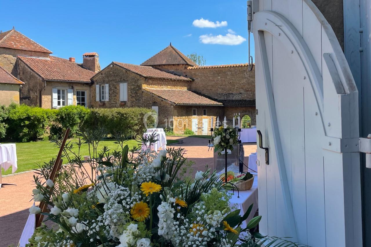 Vue d'une cour ensoleillée avec des bâtiments en pierre, des tables dressées et un bouquet de fleurs blanches et jaunes au…