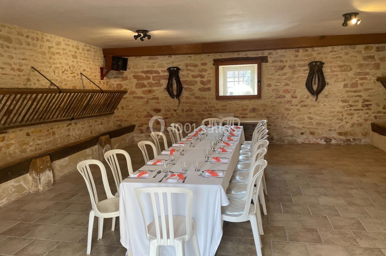 Salle en pierre avec une longue table dressée pour un repas, entourée de chaises blanches et éclairée par une fenêtre.