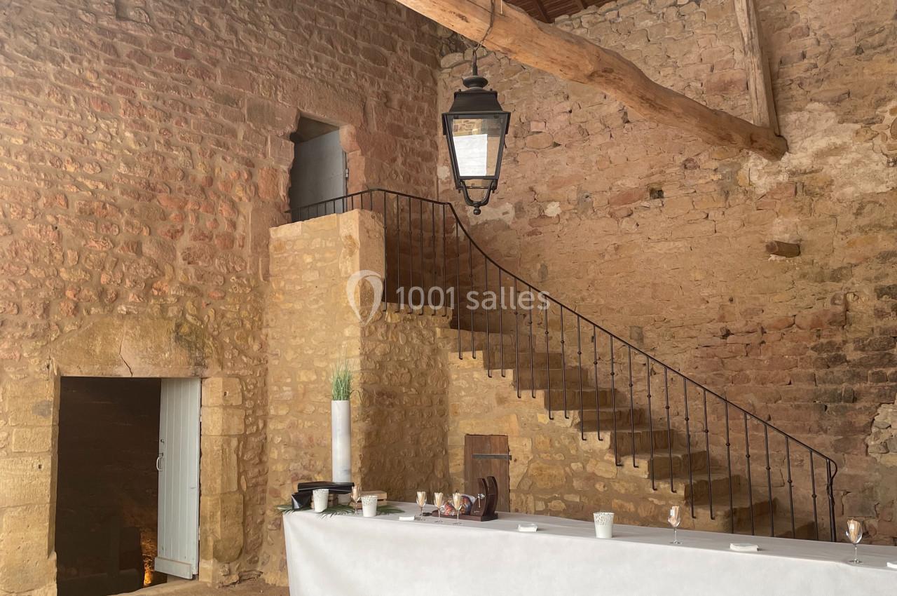 Intérieur d'une grange en pierre avec une charpente en bois, un escalier en fer forgé et une table recouverte d'une nappe…
