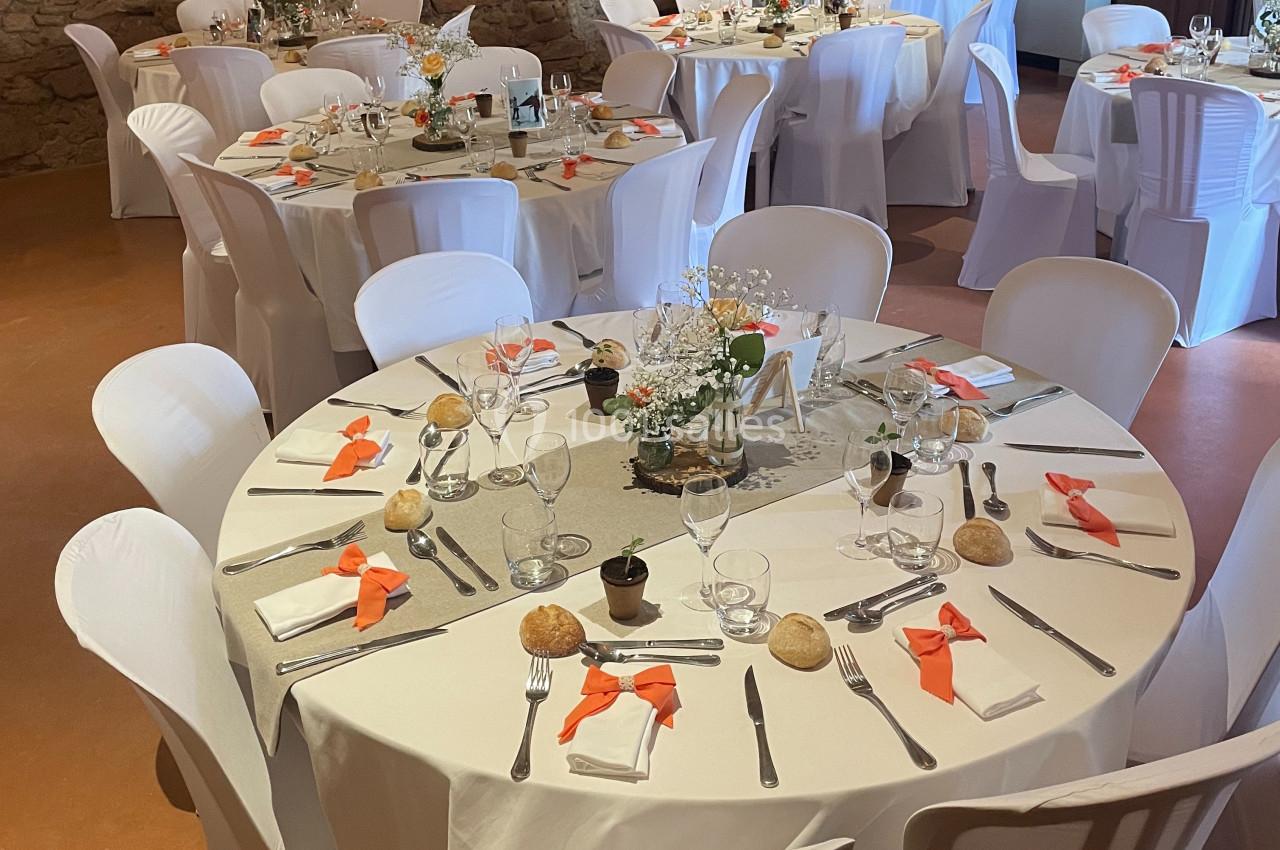 Salle décorée pour un repas, avec des tables rondes dressées, nappes blanches et touches de décoration orange.