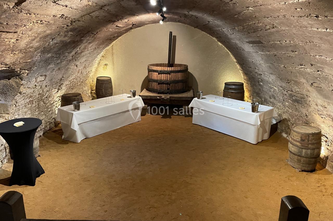 Salle voûtée en pierre avec un ancien pressoir en bois, deux tables dressées et des tonneaux en décoration.