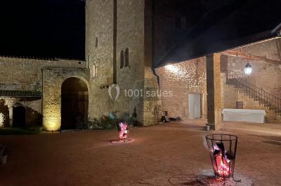 Église illuminée de nuit avec un clocher éclairé, vue depuis une allée avec des braseros allumés au premier plan.