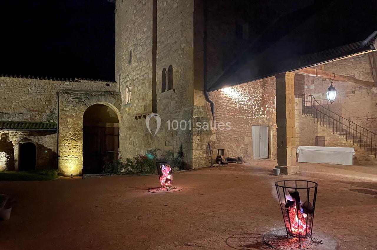 Cour d'un bâtiment en pierre éclairée la nuit, avec deux braseros allumés et un escalier en arrière-plan.