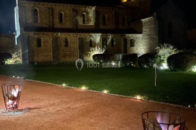 Église illuminée de nuit avec un clocher éclairé, vue depuis une allée avec des braseros allumés au premier plan.
