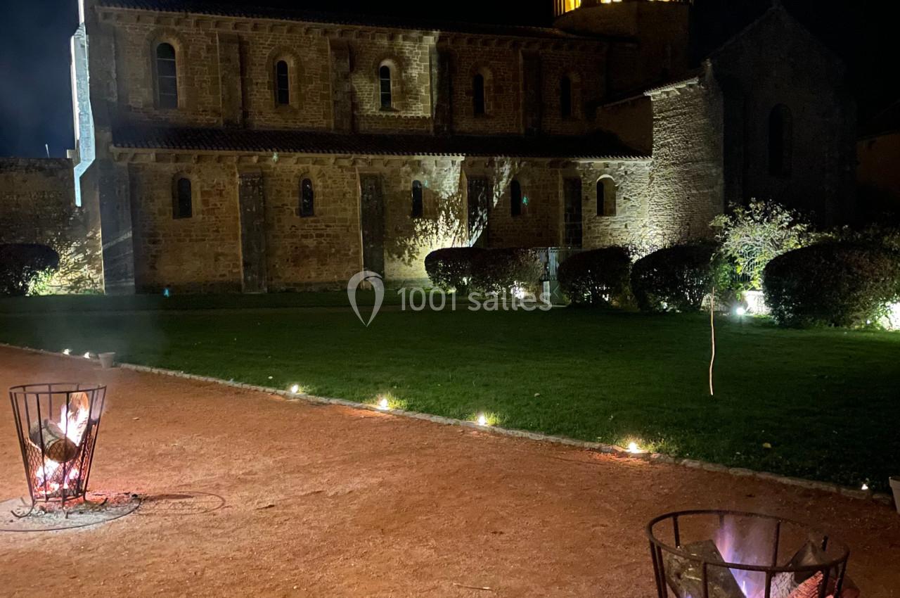 Église illuminée de nuit avec un clocher éclairé, vue depuis une allée avec des braseros allumés au premier plan.