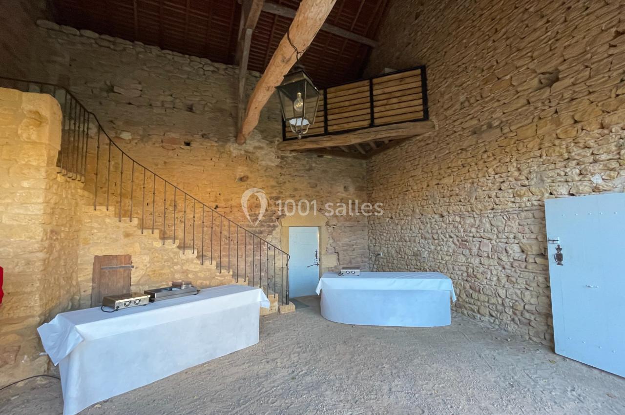 Intérieur d'une pièce en pierre avec mezzanine en bois, tables recouvertes de nappes blanches et escalier en métal.
