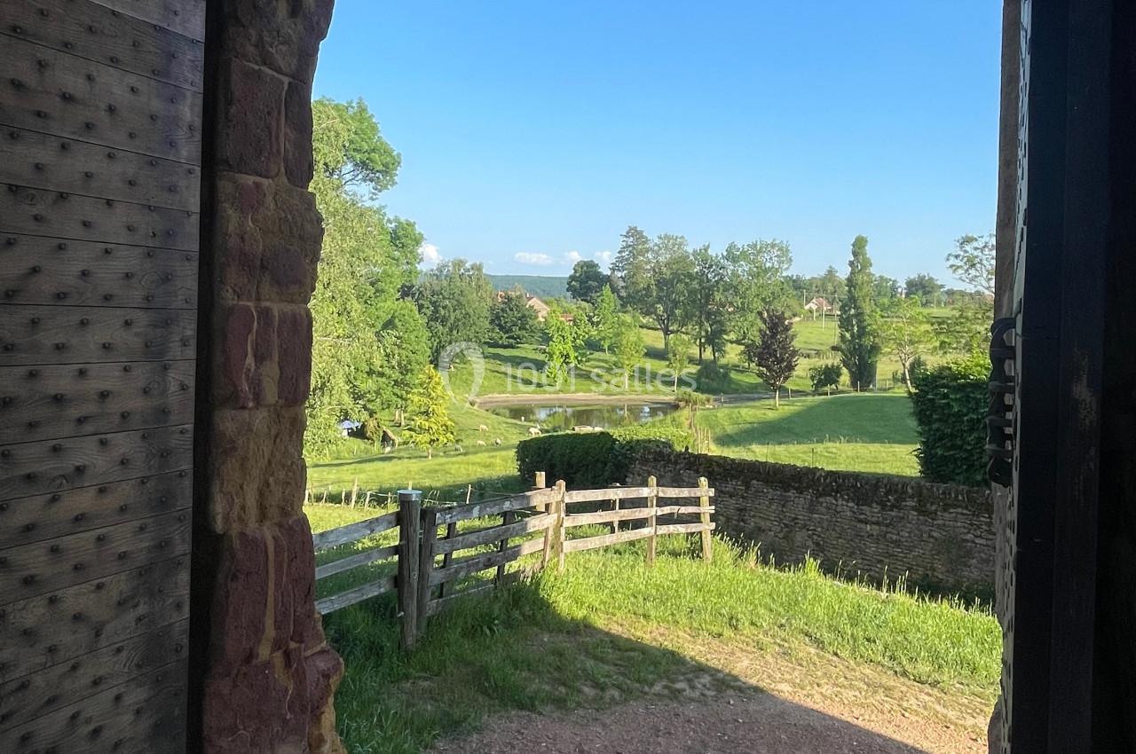 Vue sur un paysage verdoyant à travers une porte en pierre ouverte, avec une clôture en bois et un ciel dégagé.