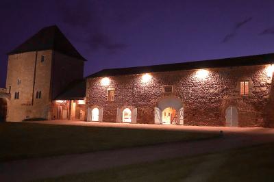 Église illuminée de nuit avec un clocher éclairé, vue depuis une allée avec des braseros allumés au premier plan.