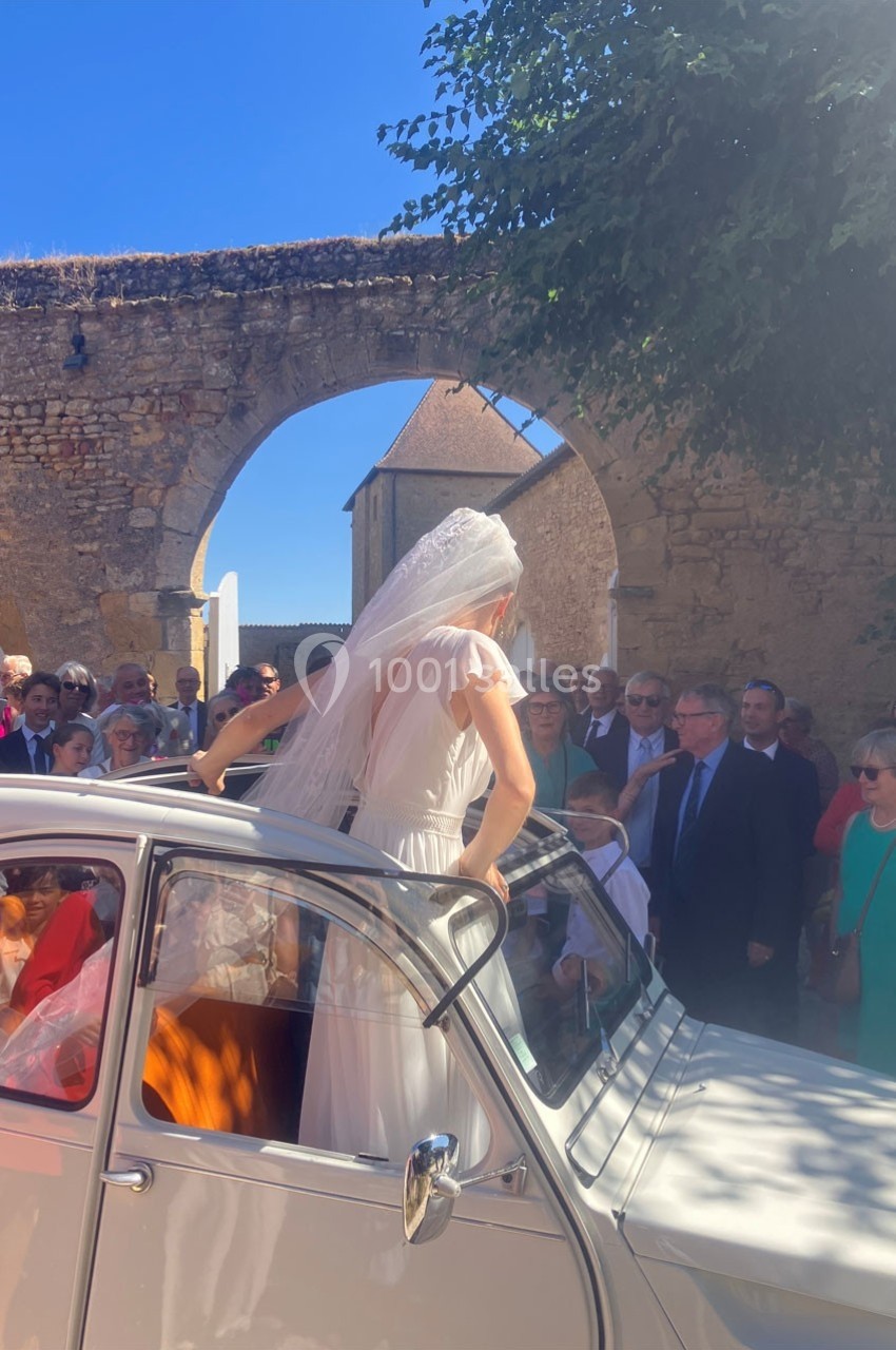 Une mariée en robe blanche et voile sort d'une voiture ancienne devant des invités rassemblés dans une cour ensoleillée.