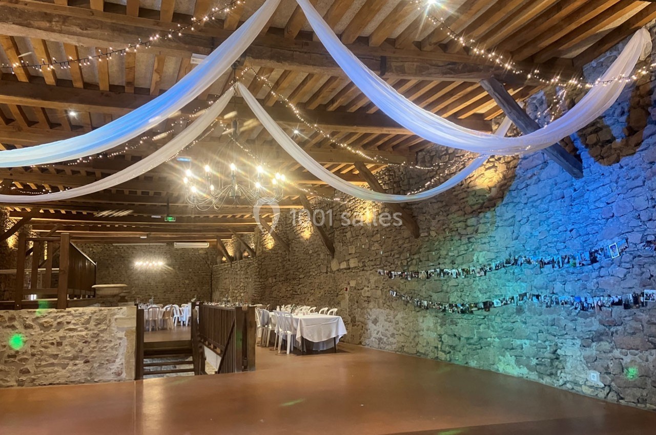 Salle en pierre avec poutres apparentes, décorée de guirlandes lumineuses, drapés blancs et tables dressées pour un…
