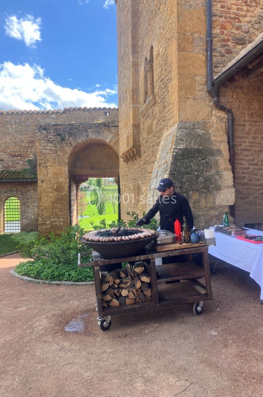 Un cuisinier prépare des saucisses sur un grand barbecue devant un bâtiment en pierre dans un cadre extérieur.