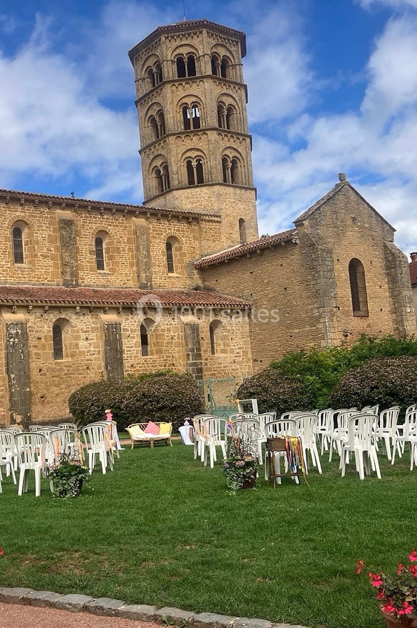 Église en pierre avec clocher roman, entourée de chaises blanches disposées sur une pelouse sous un ciel partiellement…