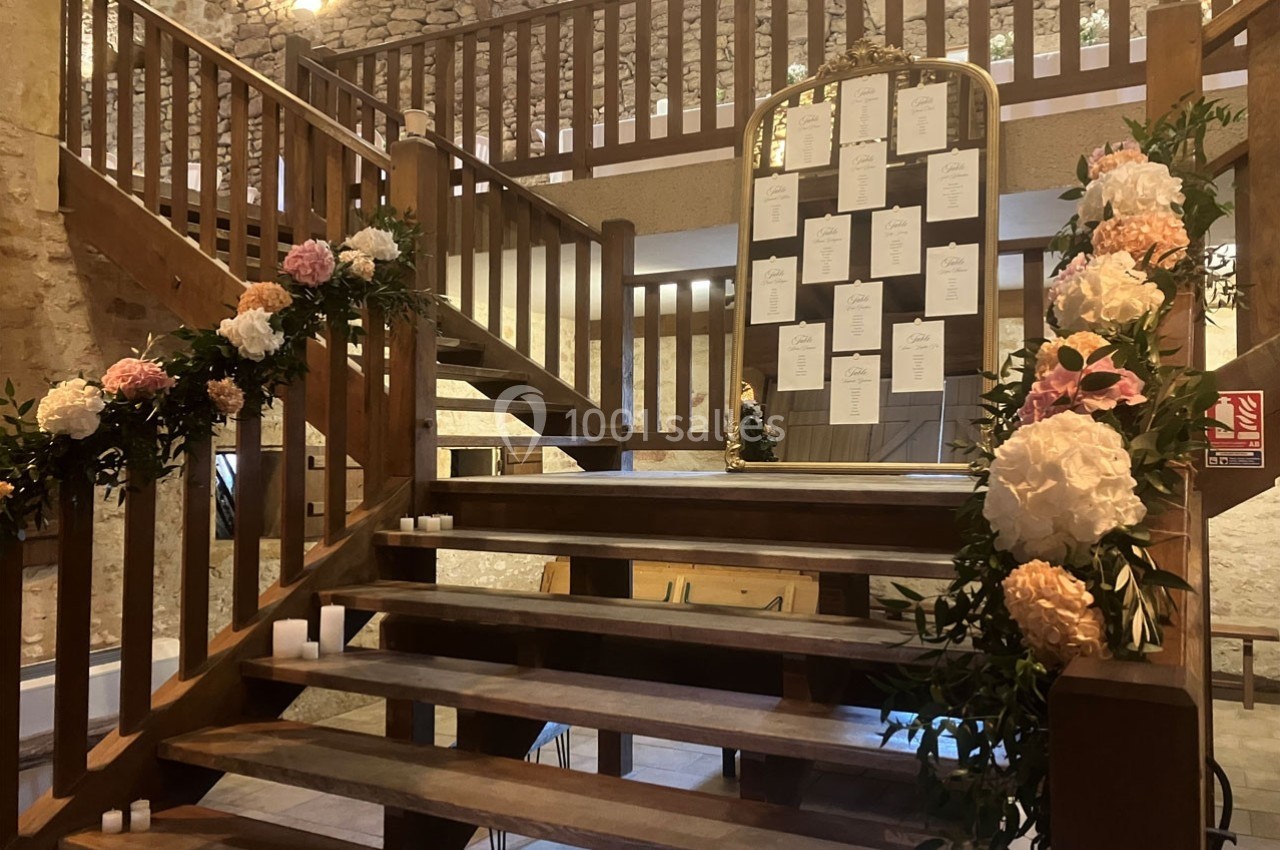 Escalier en bois décoré de fleurs et bougies, avec un tableau de plan de table placé au centre dans une salle en pierre.