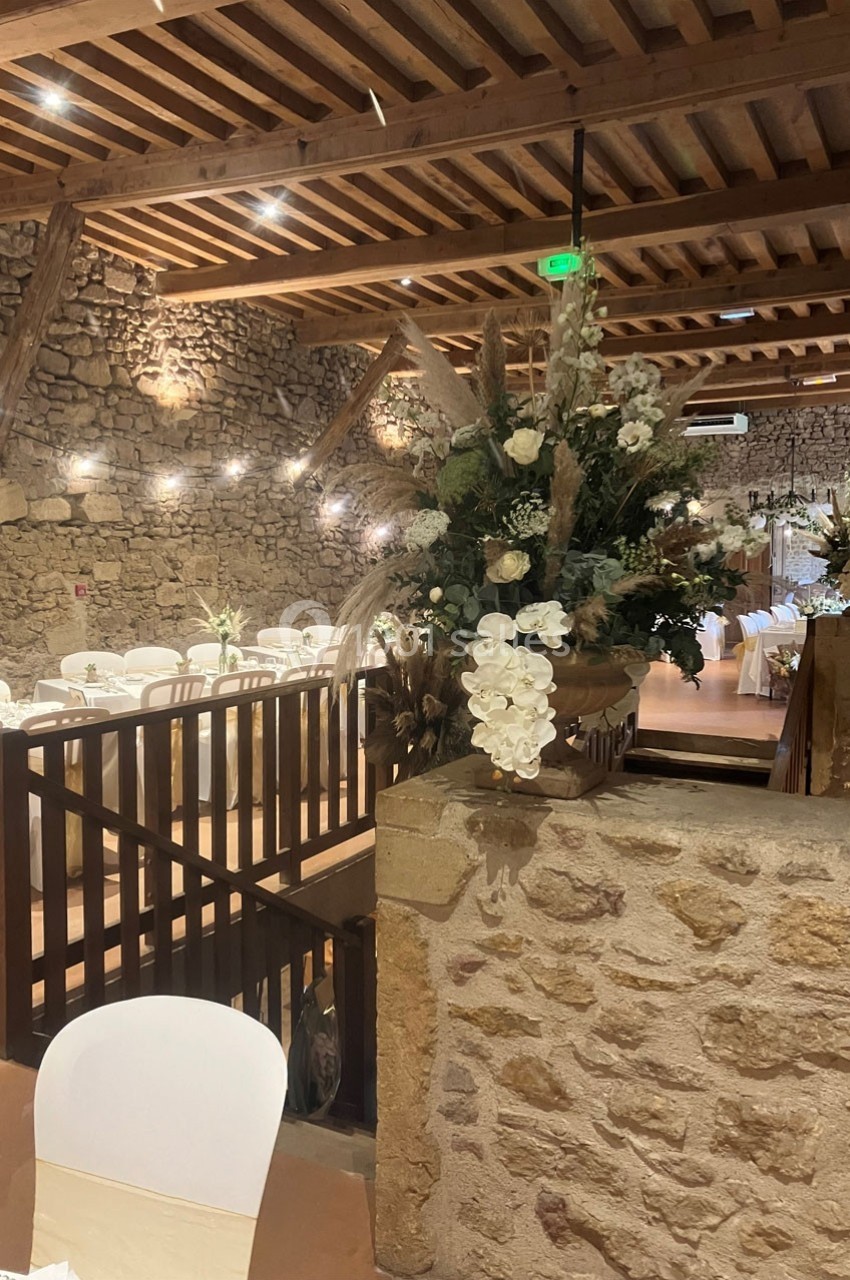 Salle de réception en pierre avec poutres apparentes, décorée de fleurs blanches et chaises habillées de housses.