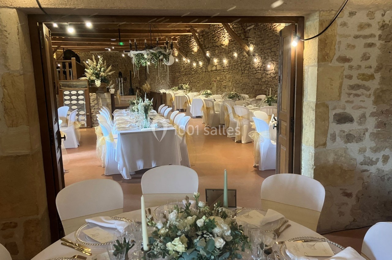Salle de réception en pierre avec tables décorées de nappes blanches, chaises ornées de rubans et centre de table floral.