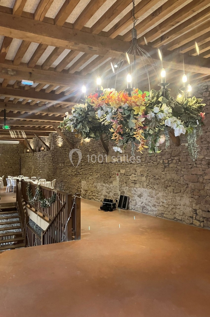 Salle en pierre avec un grand lustre décoré de fleurs suspendu au plafond en bois, tables dressées en arrière-plan.