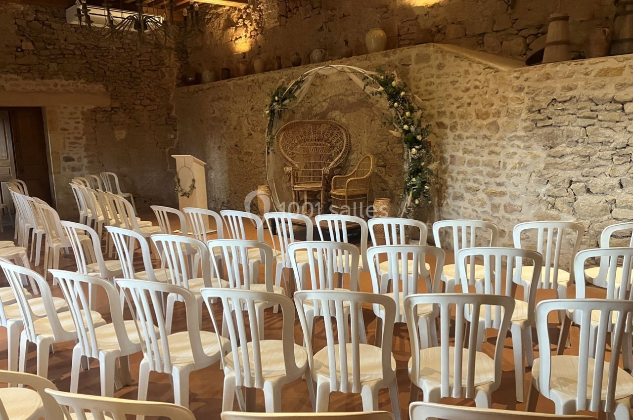 Salle en pierre aménagée pour une cérémonie, avec des chaises blanches alignées et une arche décorée de fleurs.