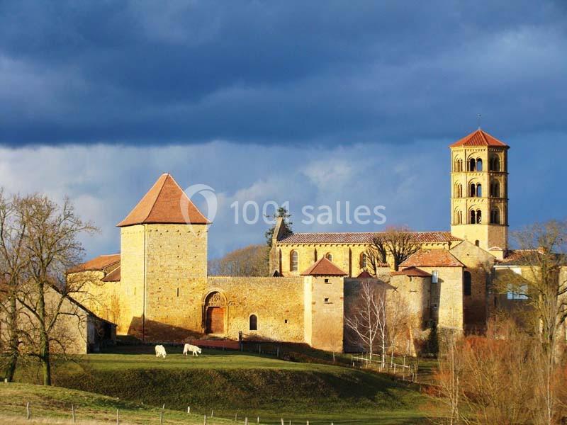 Vue d'une abbaye médiévale en pierre avec tours et clocher, entourée de prairies et d'arbres sous un ciel nuageux.