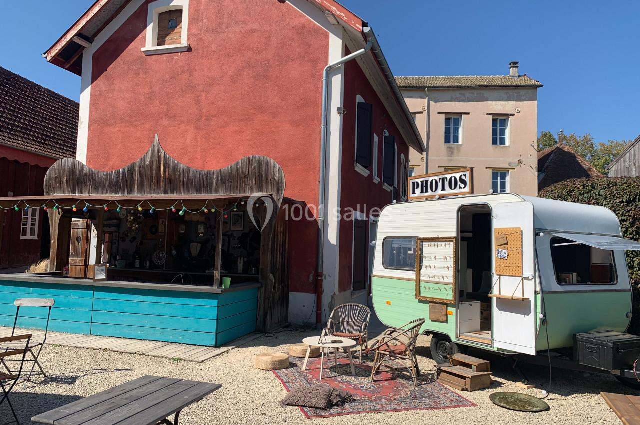 Façade d'une maison rouge avec un bar en bois et une caravane aménagée en stand photo dans une cour ensoleillée.