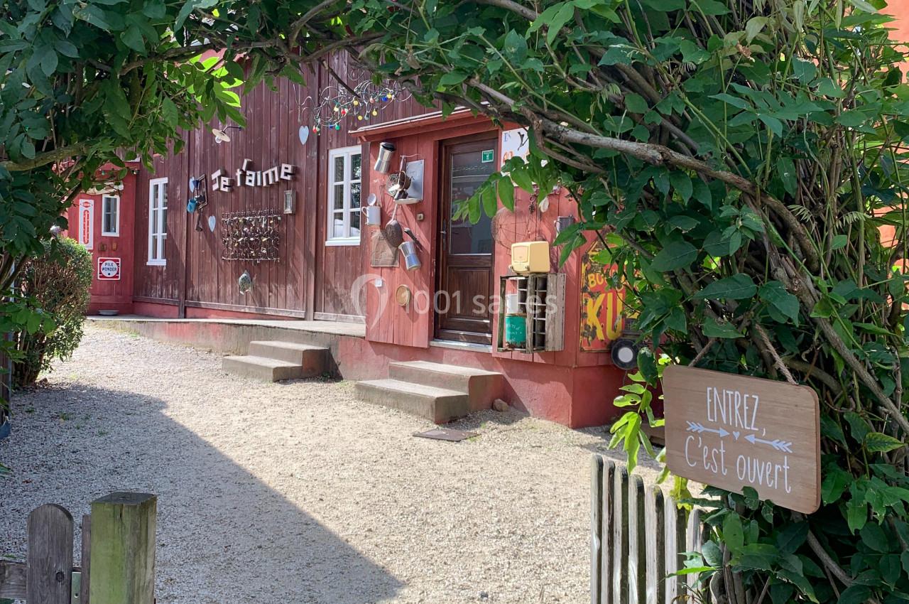 Entrée d'une ferme avec un portail entouré de verdure, un panneau ’Entrez, c'est ouvert’ et un bâtiment en bois rouge.