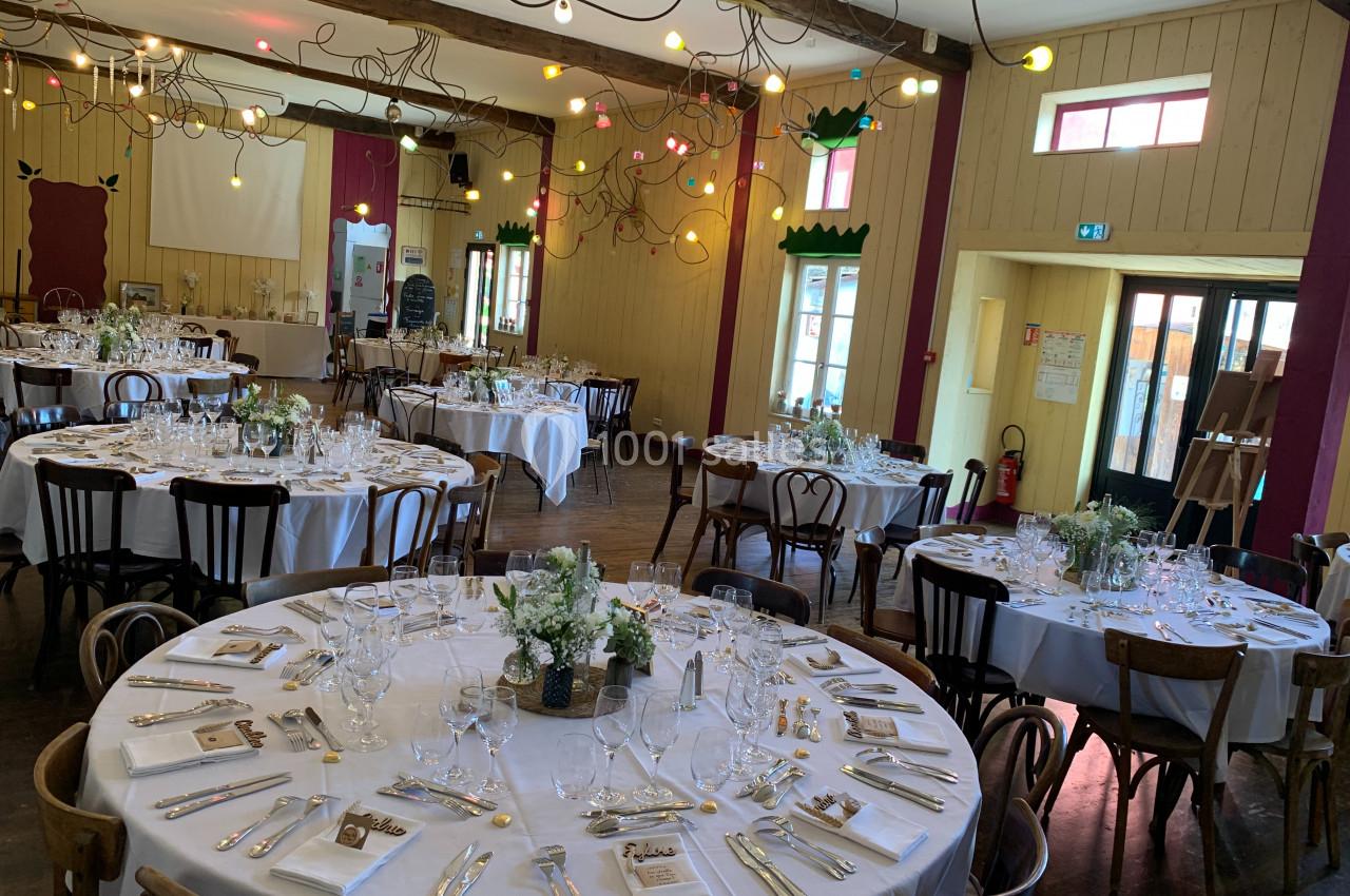 Salle décorée pour un événement avec des tables rondes dressées, nappes blanches et guirlandes lumineuses colorées.