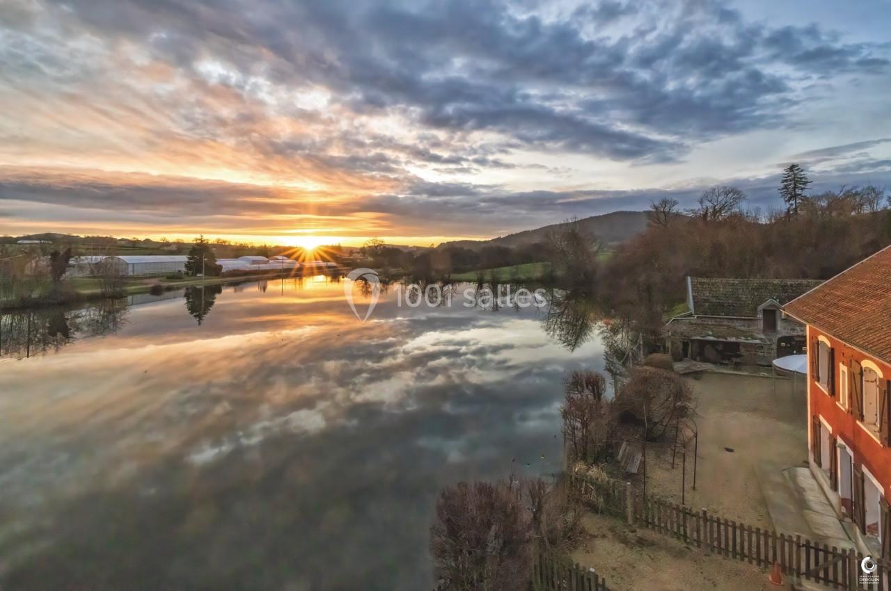 Lever de soleil sur un lac calme reflétant le ciel nuageux, entouré de végétation et de bâtiments ruraux.