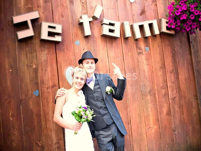 Un couple en tenue de mariage pose devant un mur en bois avec l'inscription ’Je t'aime’ et des fleurs suspendues.