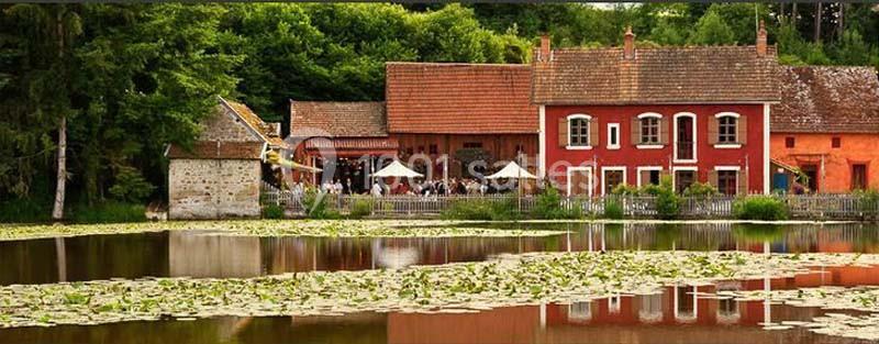 Bâtiments en briques rouges et moulin au bord d'un étang couvert de nénuphars, entourés de verdure.