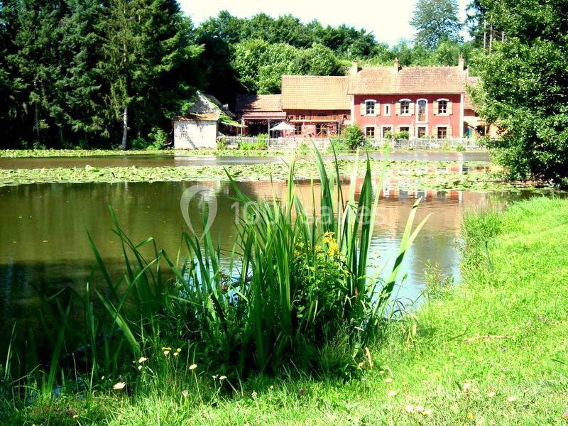 Vue d'un étang bordé de roseaux avec une maison en briques rouges et des arbres en arrière-plan.