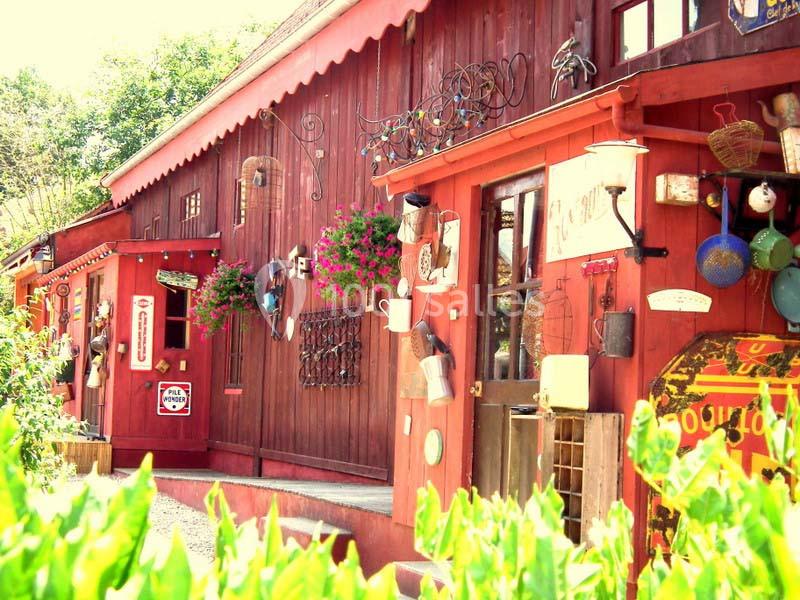 Façade d'une boutique en bois rouge décorée d'objets anciens et de fleurs suspendues, sous un ciel ensoleillé.