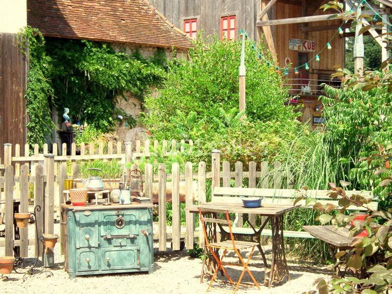 Cour extérieure rustique avec une table en bois, une cuisinière ancienne et une clôture en bois entourée de végétation.