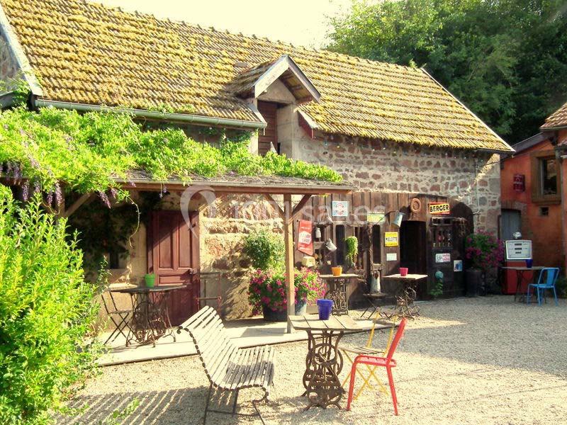 Cour pavée avec des tables, des chaises colorées et une bâtisse en pierre couverte de végétation.
