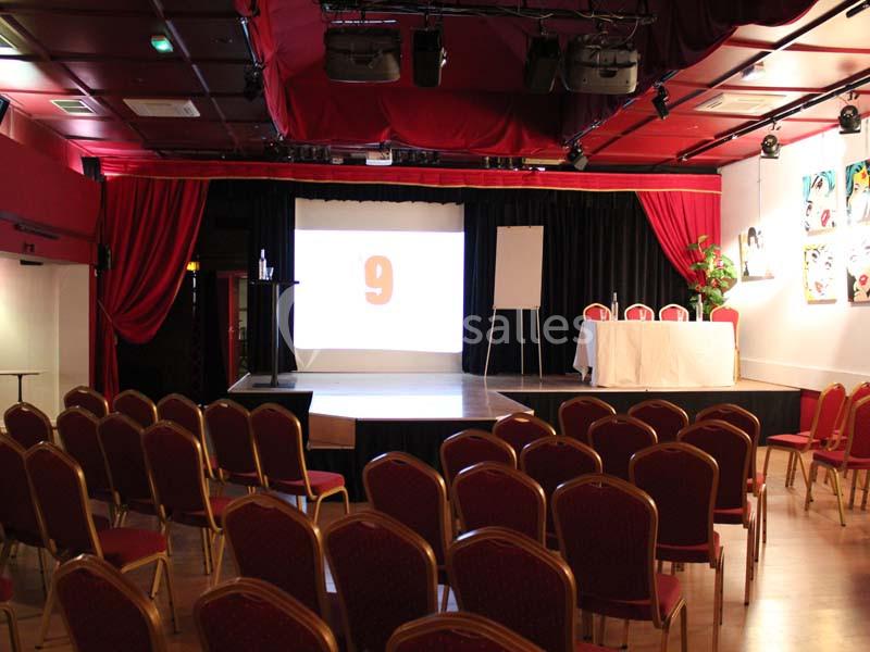 Salle de conférence avec des rangées de chaises rouges, une scène équipée d'un écran, un tableau blanc et une table.