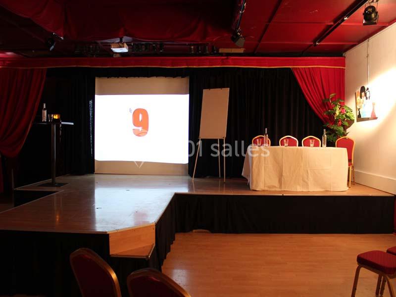 Scène avec écran de projection, tableau blanc, table dressée et chaises rouges dans une salle de conférence.
