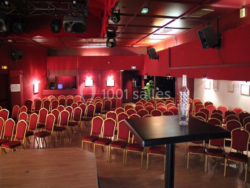 Salle de spectacle vide avec des chaises rouges alignées, éclairage tamisé et une bouteille sur une table au premier plan.