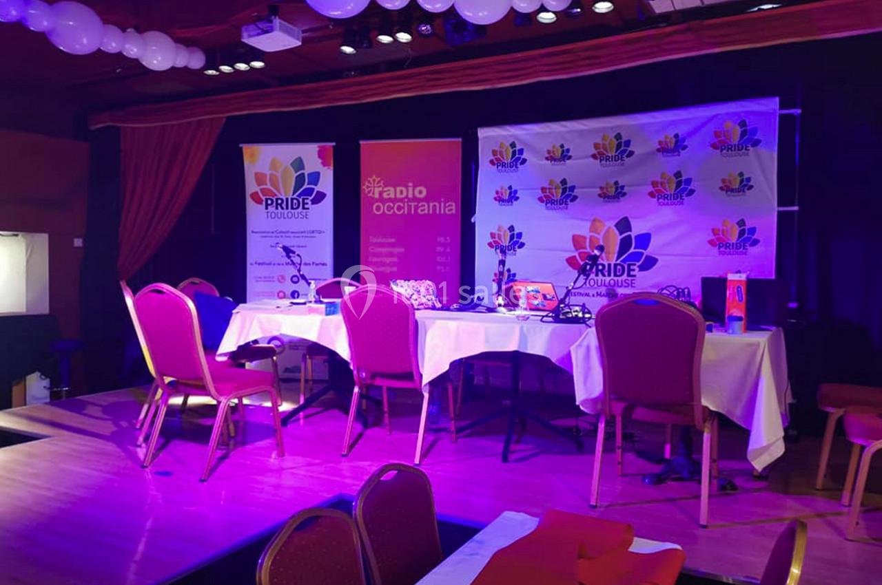 Salle décorée avec des ballons, des affiches ’Pride Toulouse’ et ’Radio Occitania’, tables et chaises disposées pour un…
