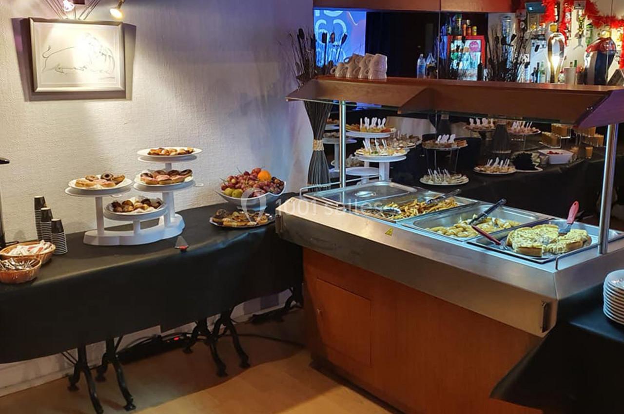 Buffet avec plats chauds, desserts variés, fruits et vaisselle disposés sur des tables dans un espace éclairé.