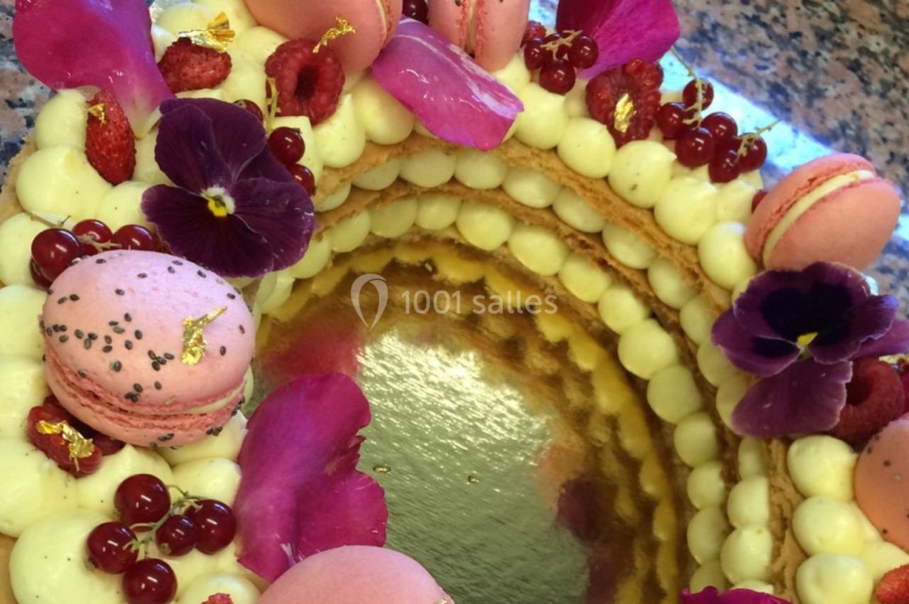 Gâteau décoré de macarons roses, fleurs comestibles, framboises et groseilles rouges sur une base de crème.