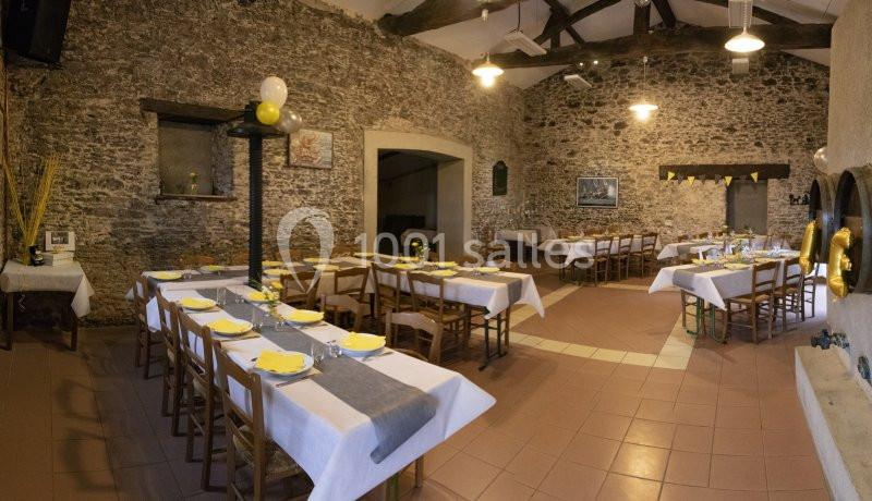 Salle en pierre aménagée pour un repas, avec tables dressées, nappes blanches et décorations jaunes et grises.
