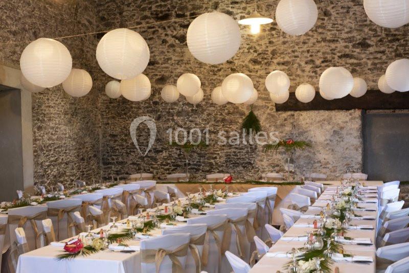 Salle décorée pour un événement, avec des tables alignées, nappes blanches, chaises ornées et lanternes suspendues.