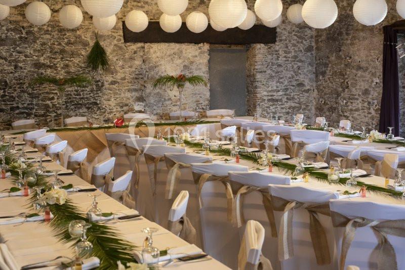 Salle de réception décorée avec des tables alignées, nappes blanches, chaises ornées et lanternes suspendues.
