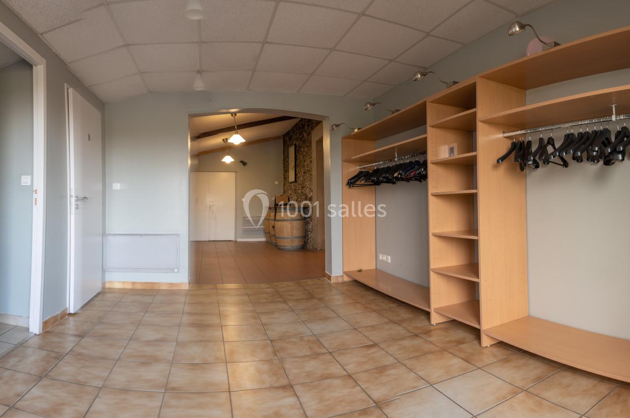 Vestiaire avec étagères en bois et portants à cintres, sol carrelé et vue sur un couloir menant à une pièce avec un tonneau.