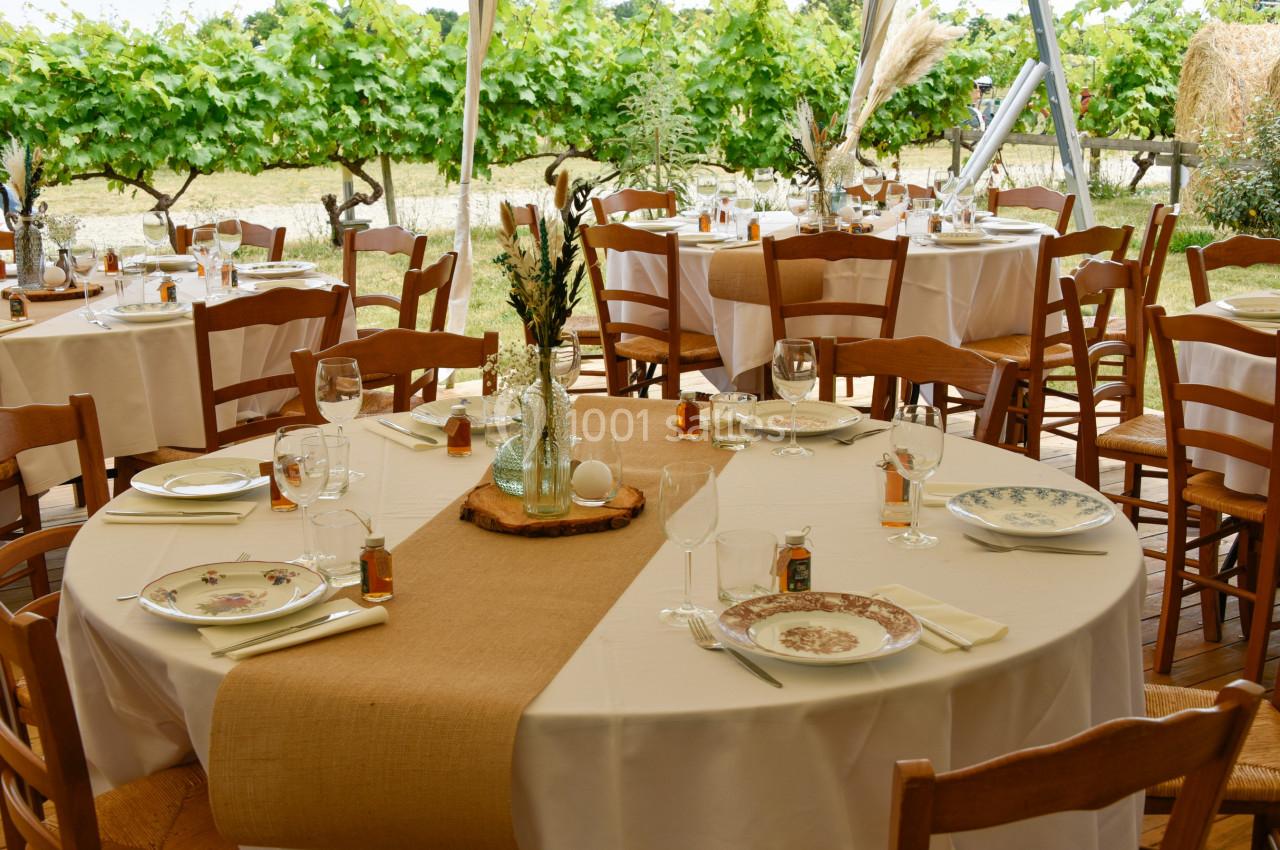 Tables rondes dressées avec nappes blanches, vaisselle élégante et décor champêtre, en extérieur près de vignes.