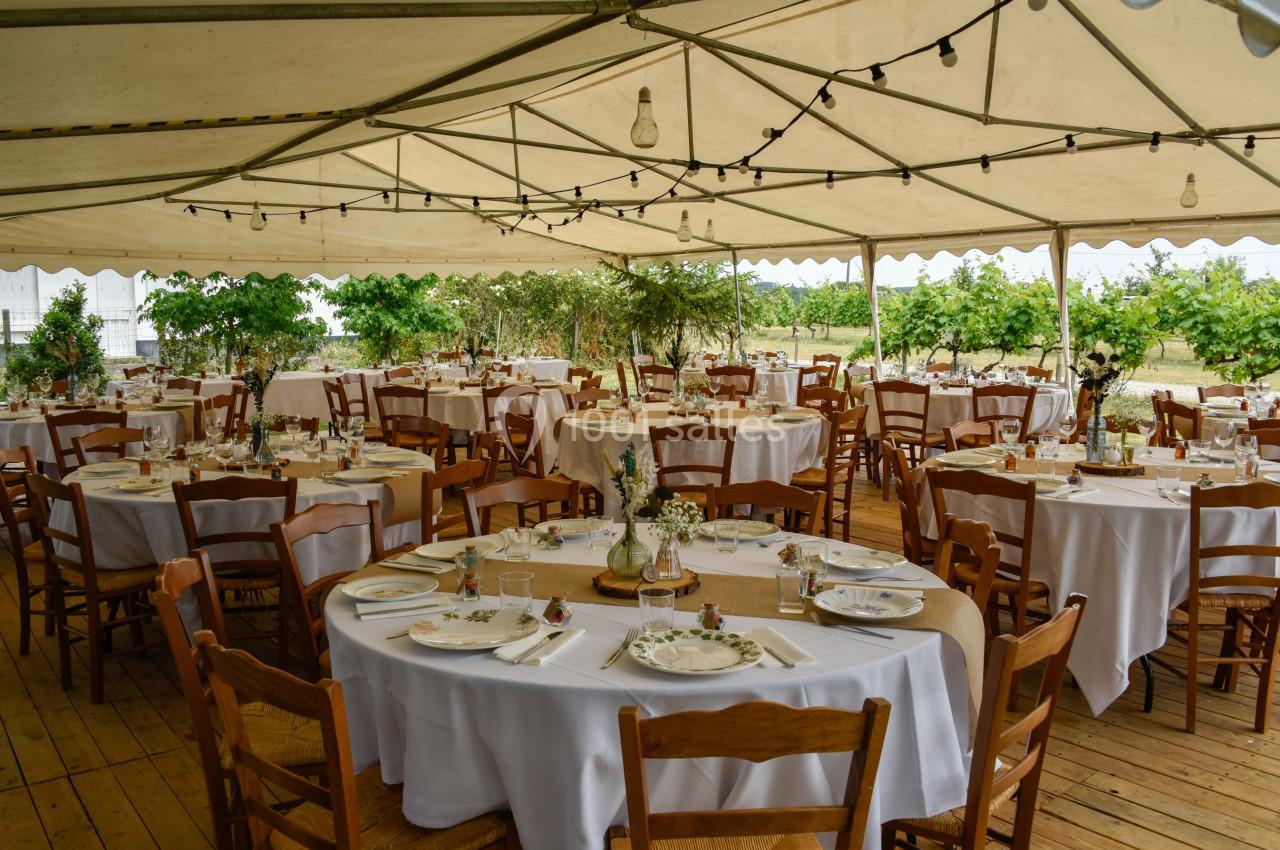 Salle de réception en plein air sous une tente, avec tables rondes dressées et décorées pour un événement.