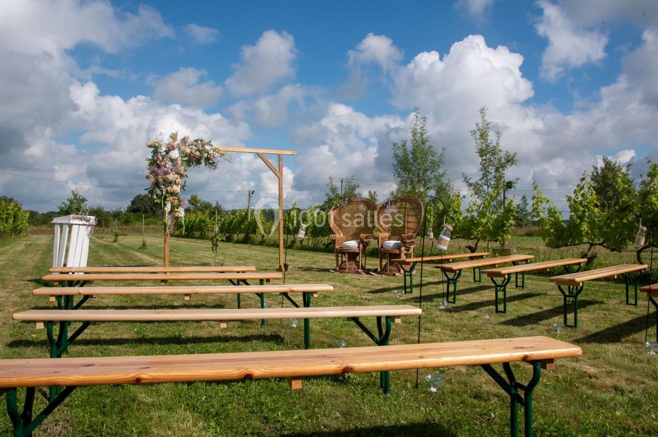 Bancs en bois disposés en extérieur face à une arche fleurie, dans un cadre champêtre sous un ciel partiellement nuageux.