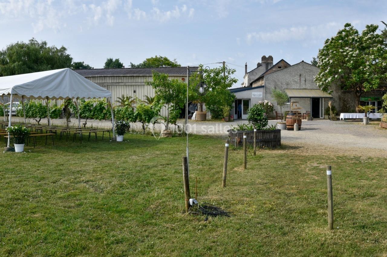 Vue d'un espace extérieur avec une tente blanche, des tables, des chaises et des bâtiments entourés de verdure.