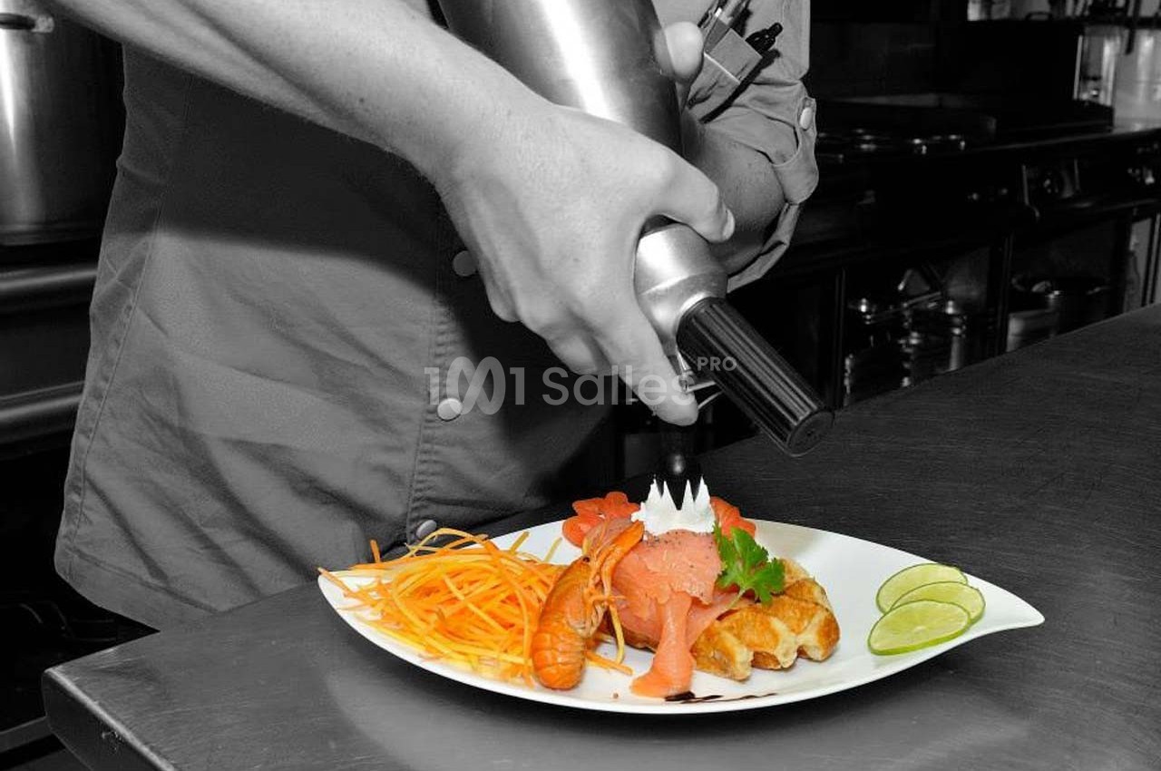 Un chef décore une assiette de saumon, crevettes et légumes avec une mousse à l'aide d'un siphon en cuisine.