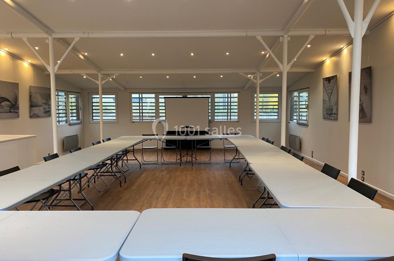 Salle de réunion lumineuse avec tables disposées en U, chaises noires et écran de projection au fond.