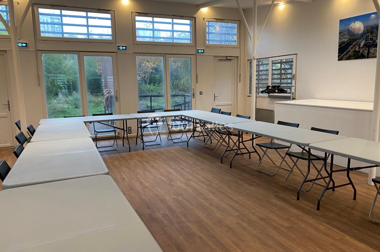 Salle lumineuse avec tables en U, chaises pliantes, parquet au sol et grandes baies vitrées donnant sur un espace vert.