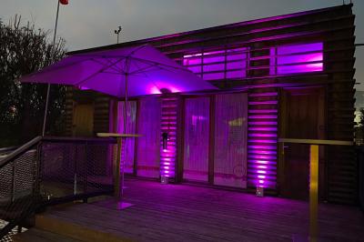 Façade en bois éclairée par des lumières violettes, avec une terrasse en bois et un grand parasol.